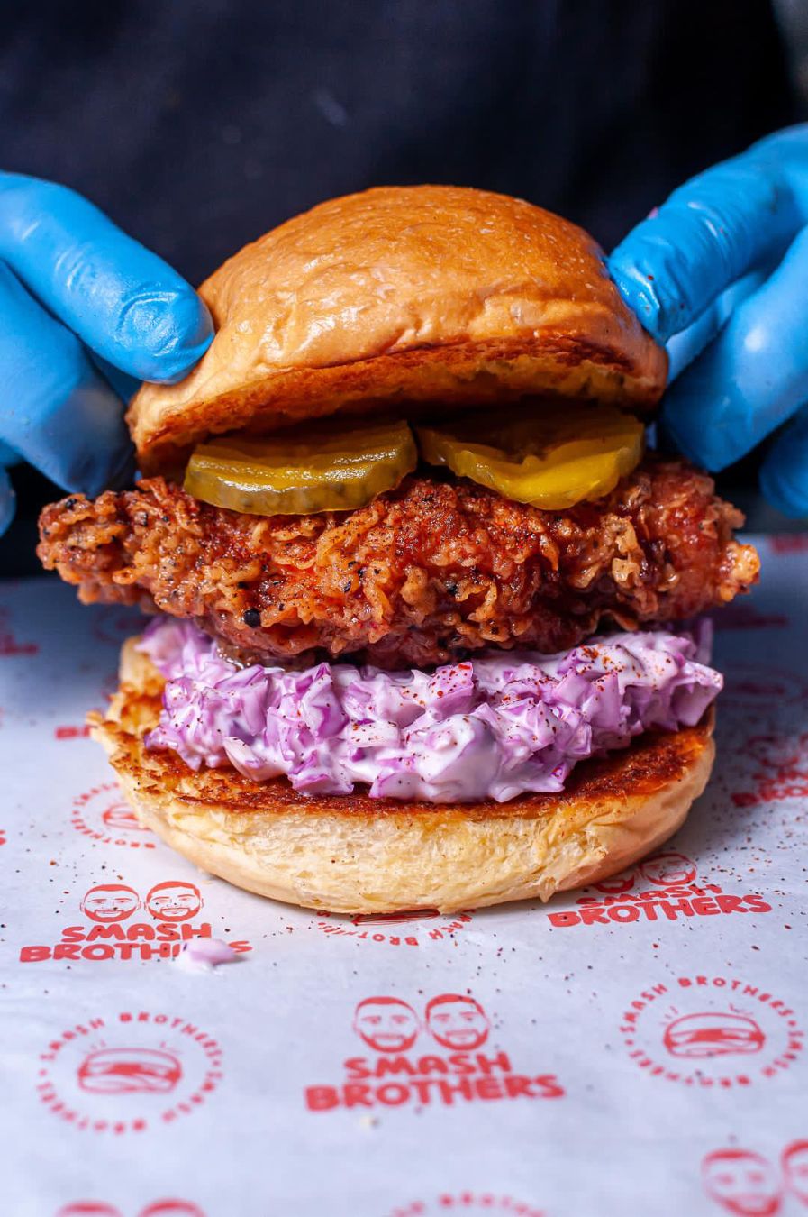 Crispy chicken sandwich with purple slaw, held by hands in blue gloves